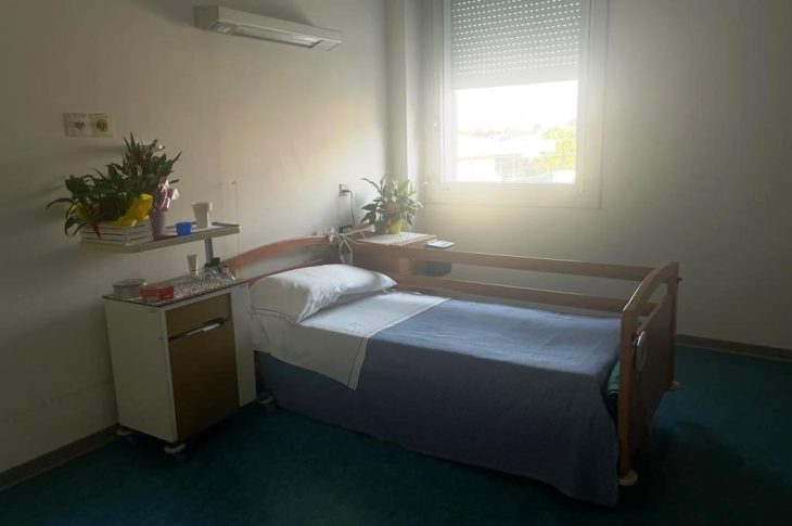 Una stanza singola della casa di riposo: c'è un letto con a fianco la finestra e due comodini con alcune piante in vaso